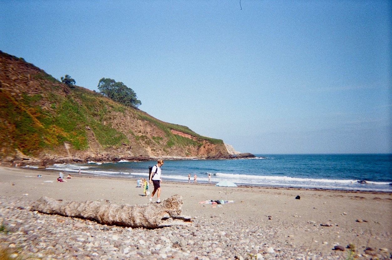 A scenic beach landscape with a sandy shore, pebbled foreground, blue ocean, and a grassy rocky cliff under a clear sky. A few small people are visible near the water and on the beach.