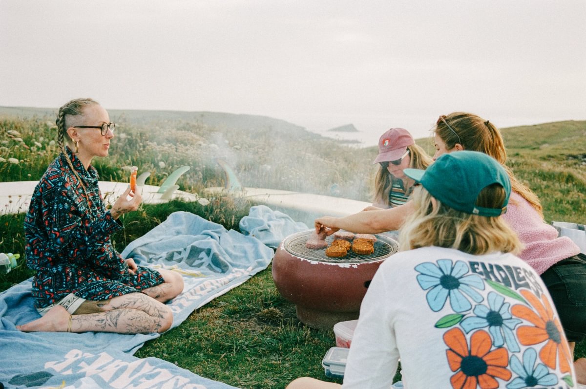 A group of friends enjoys an outdoor picnic and grilling session on a grassy hillside under an overcast sky. One person wears a colorful geometric long-sleeve top, while another in the foreground wears a white t-shirt with a large blue, orange, and green flower graphic on the back.