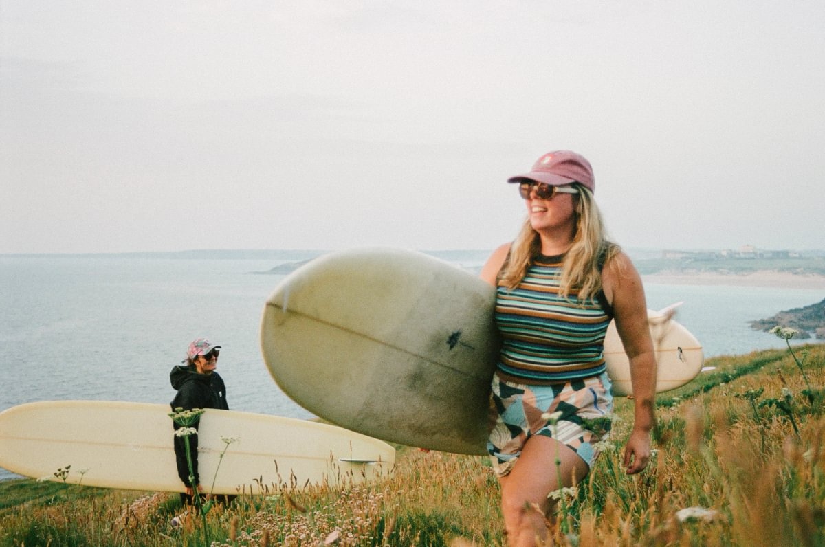 A woman carrying a surfboard walks along a grassy coastal path, wearing a pink cap, sunglasses, a striped tank top, and a patterned skirt. Another person with a surfboard stands in the background near the ocean.
