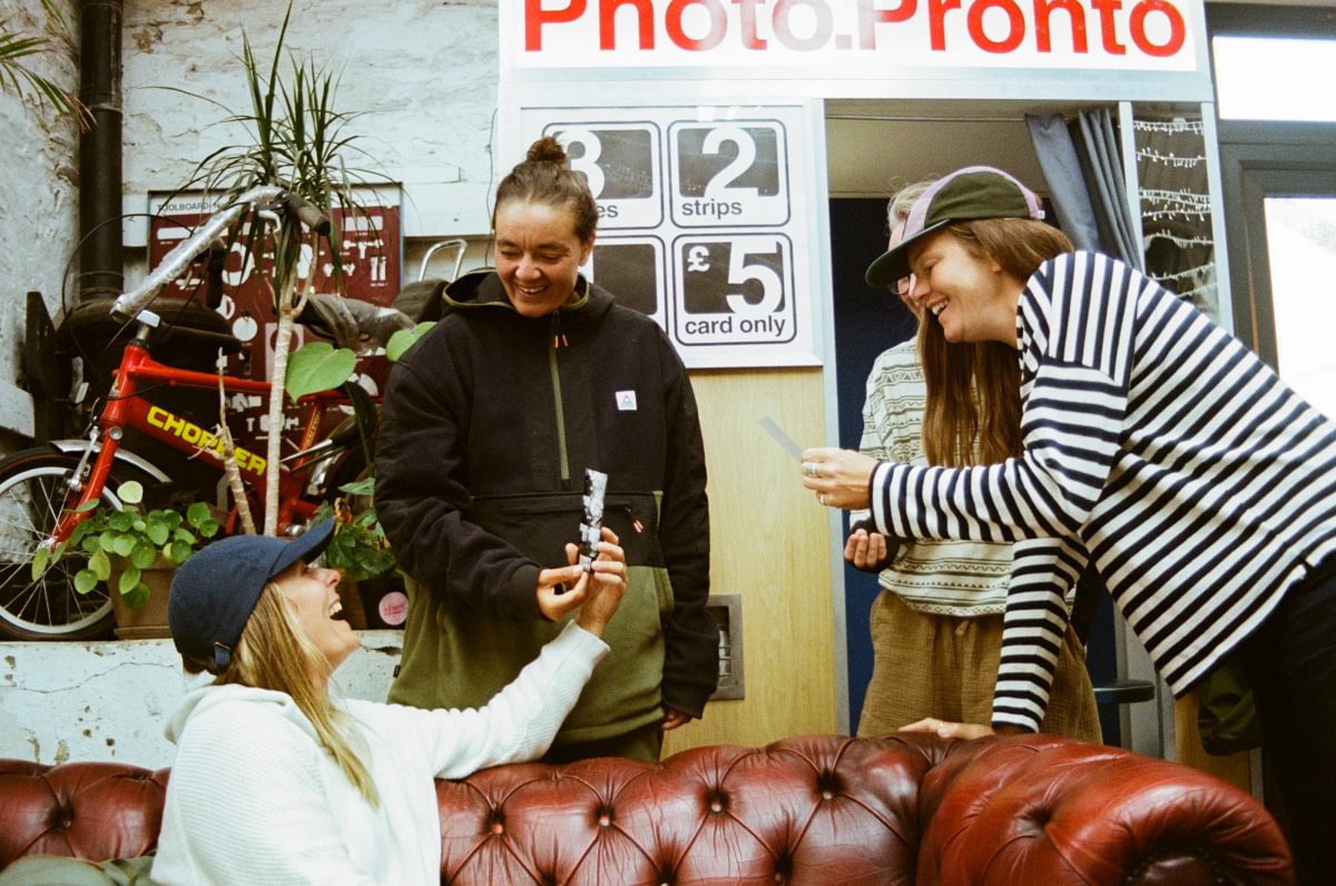 Three women are gathered in a shop setting, interacting with a photo booth. A red leather tufted sofa is in the foreground, and a bicycle is visible on the left.