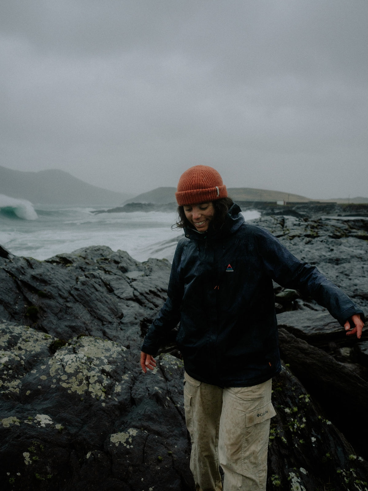A person standing on dark volcanic rocks by the ocean under an overcast sky, wearing a dark navy jacket, light beige pants, and a rust-orange knit beanie.