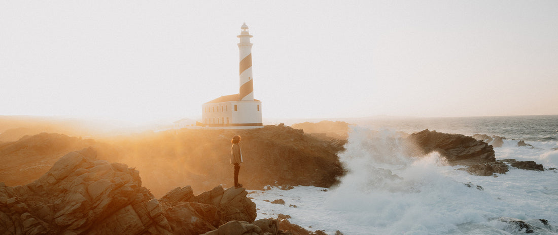 A woman stands on a cliff in golden light, lighthouse behind