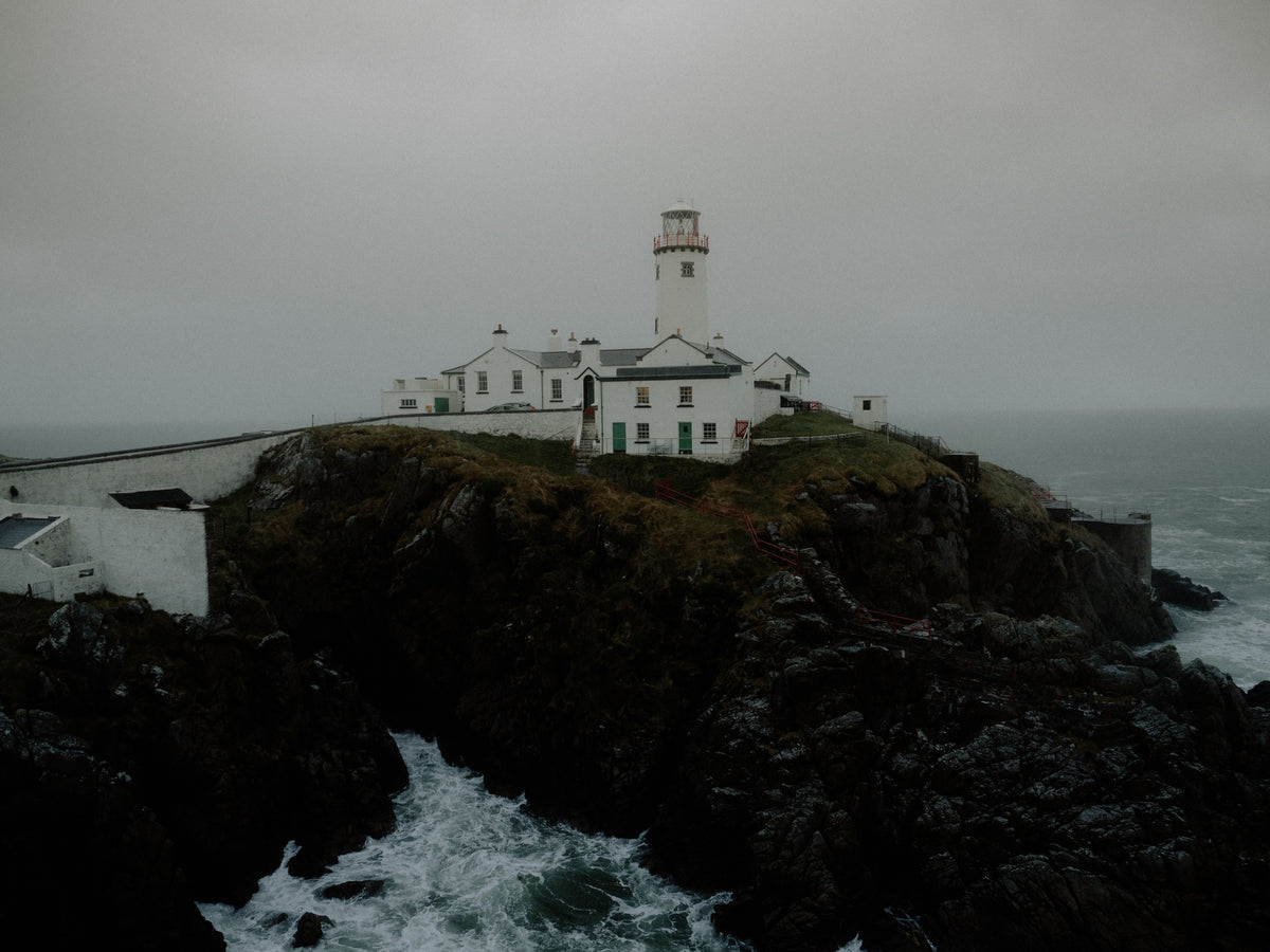 A lighthouse and white buildings sit on a rocky coastal cliff with dark rocks and churning ocean water under an overcast gray sky.