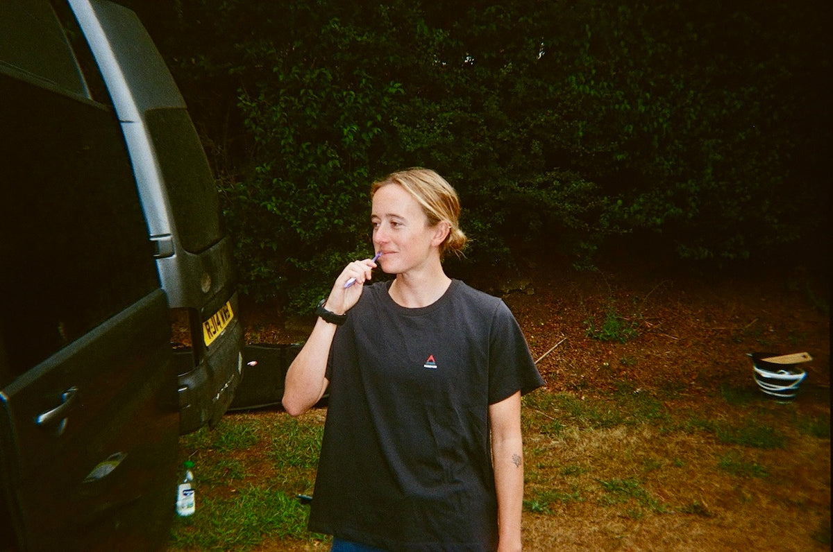 A woman wearing a black short-sleeve t-shirt with a small red and blue logo on the chest stands outdoors next to a dark vehicle at night, holding a phone to her ear. The background is dark with trees and grass.