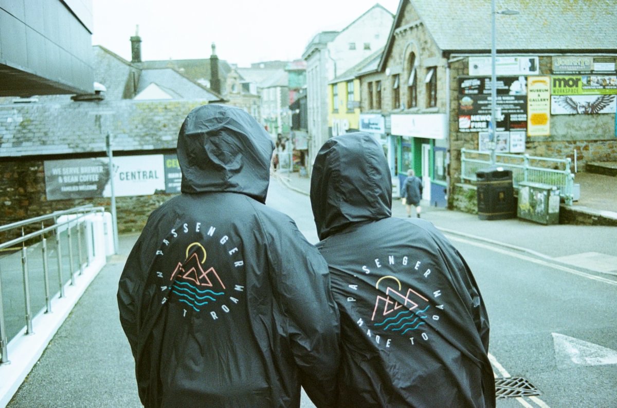 Two people wearing black hooded change robes with a mountain logo and the words 'Passenger Made To Roam' on the back, standing on a street in a town setting.