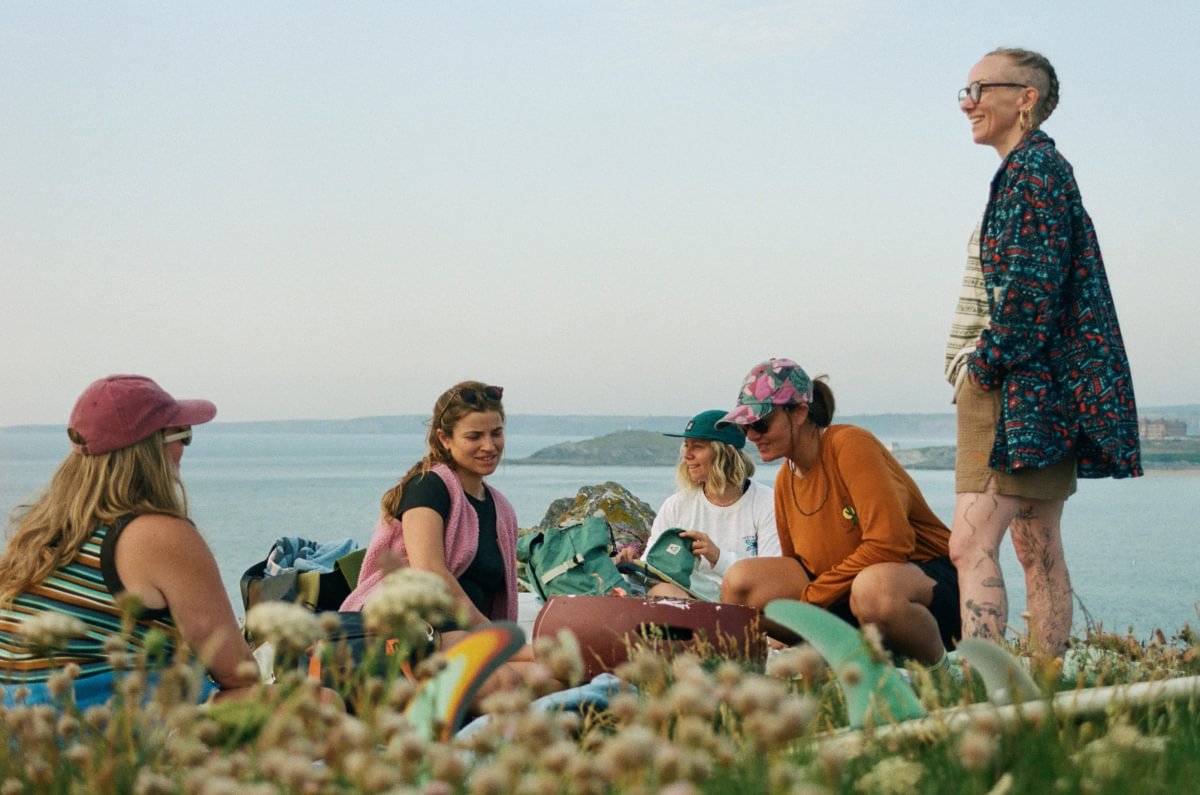 A group of five women are gathered outdoors on a grassy cliff overlooking the sea under a clear sky. One woman stands on the right wearing a dark geometric patterned overshirt, beige shorts, and glasses, while the others sit in the foreground wearing casual beachwear.